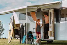 Couple relaxing by a motorhome with awning, surfboard, and camping gear outdoors