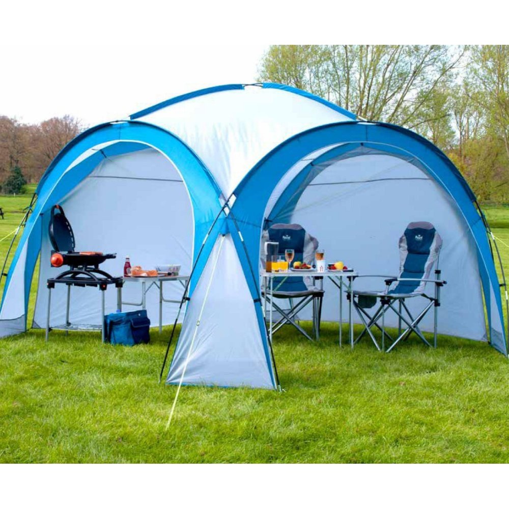 Blue and white camping shelter tent with outdoor chairs, tables, and a portable grill set up on grass.